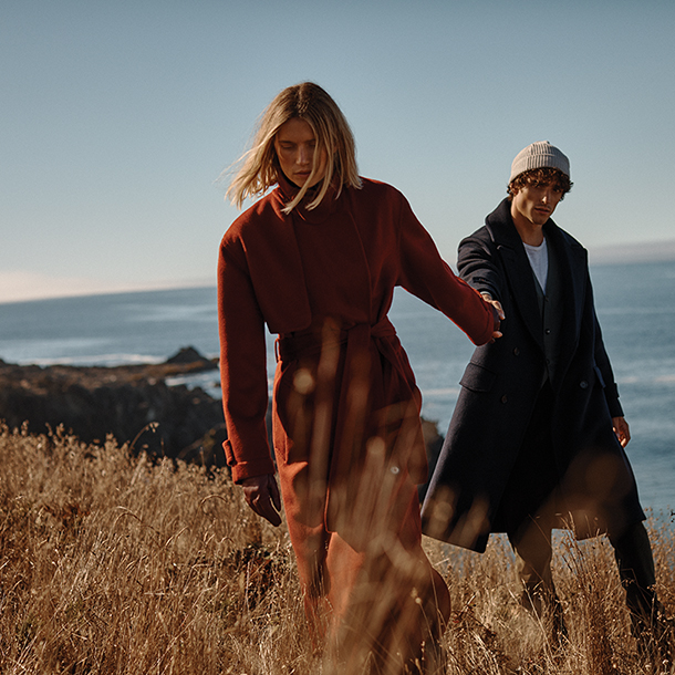 A couple, dressed in long coats, holds hands while strolling along a clifftop path overlooking the ocean.